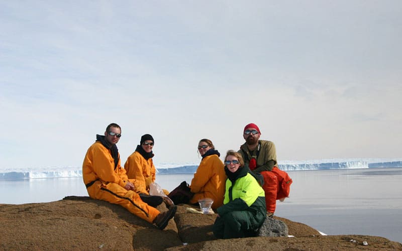 Five people in the foreground sitting on brown ground with large ice sheets behind them.