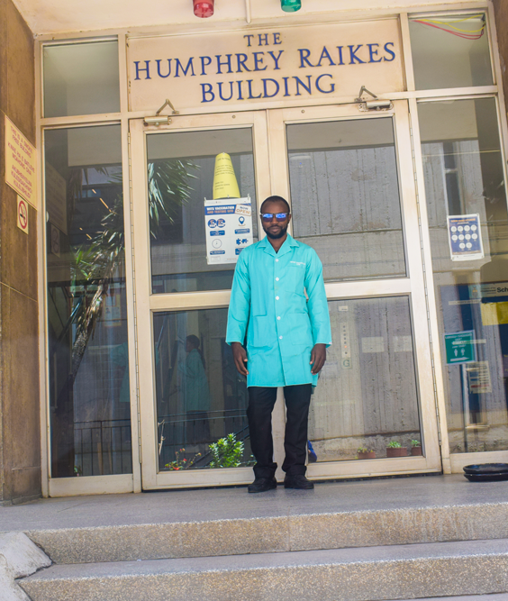 Image of Jean Baptiste Fankam Fankam standing at the entrance of the Humphrey Raikes Building, and he is dressed in a lab coat and lab eye glasses.
